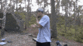 a man in a hat is standing in the woods with his hands in his pockets .
