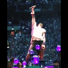 a man in a white shirt is holding a microphone in front of a crowd with purple lights behind him