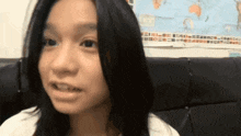 a young girl with long black hair is sitting on a couch with a map of the world behind her .