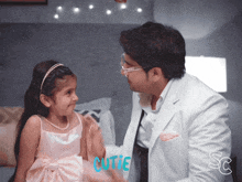 a little girl in a pink dress talks to a man in a white suit with the word cutie in blue