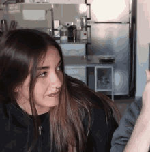 a woman in a black hoodie is sitting in a kitchen with a fridge in the background .