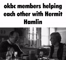 a black and white photo of a man and woman sitting at a table with okbc members helping each other with hermit hamlin