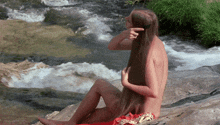 a naked woman is sitting on a rock by a stream brushing her hair