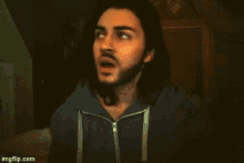 a man with long hair and a beard is wearing a blue hoodie and adjusting his hair .