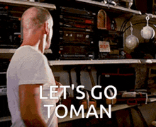 a bald man in a white shirt is standing in front of a shelf with the words let 's go toman written on it