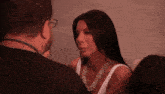 a woman wearing a white tank top and a necklace talks to a man wearing glasses