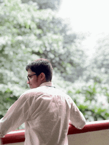 a man wearing glasses and a white shirt stands on a balcony with trees in the background
