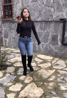 a woman in a black top and blue jeans is standing on a stone walkway holding a cell phone