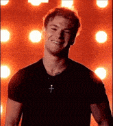 a young man wearing a black shirt and a cross necklace is smiling in front of a red background .