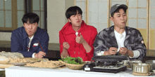three young men are sitting at a table eating food with chopsticks and one of them is wearing a nike jacket