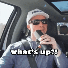 a man wearing sunglasses and a friday hat is holding a microphone and says what 's up