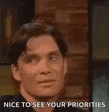 a man is sitting in front of a brick wall with the words `` nice to see your priorities '' written below him .