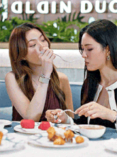 two women are sitting at a table with plates of food in front of a sign that says plain duc
