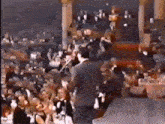 a man in a suit is standing on a stage in front of a crowd of people applauding .