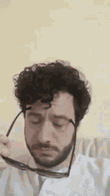 a man with curly hair and a beard is holding a pair of sunglasses over his eyes .