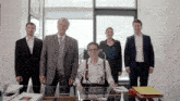 a man in suspenders sits at a desk in front of a group of men