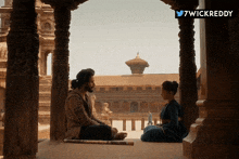 a man and a woman are sitting in front of a building with the twitter username 7wickreddy above them