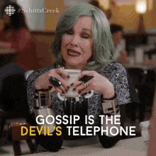 a woman with green hair is sitting at a table holding a cup of coffee and saying gossip is the devil 's telephone