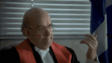 a man wearing glasses and a red robe is sitting in front of a flag