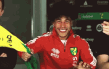 a man wearing a red errea sweatshirt smiles while holding a yellow flag
