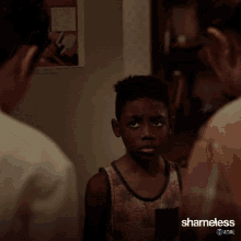 a young boy stands in front of a group of people with shameless written on the bottom right