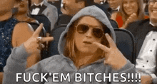 a woman in a hoodie and sunglasses is sitting in a crowd of people and making a peace sign .