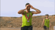 a man in a green tank top is covering his eyes with his hands while another man stands behind him .