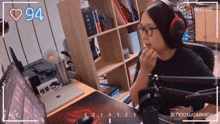a woman wearing headphones and glasses is sitting at a desk with a monitor and a heart 94 in the background