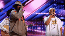 a man singing into a microphone while another man plays a guitar in front of a nbc logo