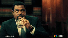 a man in a suit and tie is holding a glass of whiskey with a nbc logo in the corner