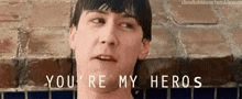 a man is standing in front of a brick wall with the words `` you 're my heros '' written on his face .