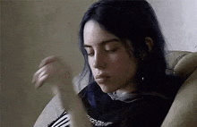 billie eilish is sitting on a couch with her eyes closed and her hand in her mouth .