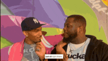 a man wearing a ny hat talks to another man wearing a shirt that says sucka