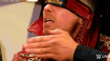 a wrestler wearing a red and gold outfit and a blindfold is holding his hands to his face .