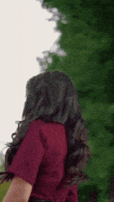 a woman in a red shirt is standing in front of a tree and looking at the camera .