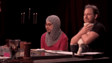 two men are sitting at a table with their arms crossed and one of them is wearing a chain mail helmet .