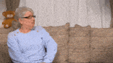 an older woman wearing glasses sits on a couch with a teddy bear in the background