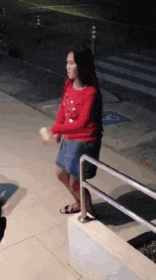 a girl in a red sweater and blue skirt stands on a sidewalk