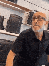 a bald man wearing glasses and a black shirt stands in front of a display of suitcases