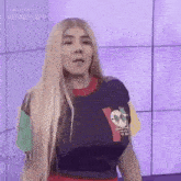 a woman with long blonde hair is standing in front of a purple wall wearing a blue shirt .