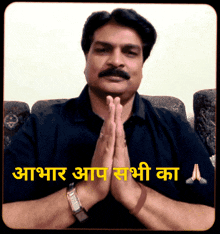 a man with a mustache is praying with his hands folded in front of a sign that says " आप सभी का "