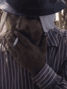 a man wearing a hat and a striped shirt smoking a cigarette .