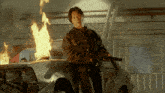 a man is holding a gun in front of a car that is on fire