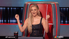 a woman sitting in a chair with her arms in the air and a nbc logo in the background