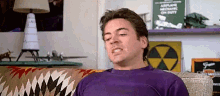 a man in a purple shirt is sitting on a couch and making a funny face .