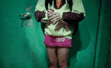 a girl wearing a yellow sweater and pink shorts stands in front of a green wall