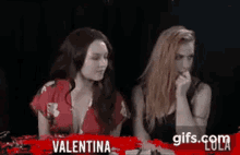 two women are sitting next to each other in a dark room . one of the women is wearing a red shirt .