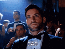 a man in a tuxedo and bow tie is standing in front of a crowd of people and saying dad .