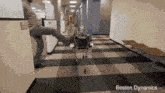 a man is standing next to a robot in a hallway with boston dynamics written on the bottom .