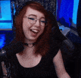 a woman wearing glasses and a choker smiles in front of a microphone